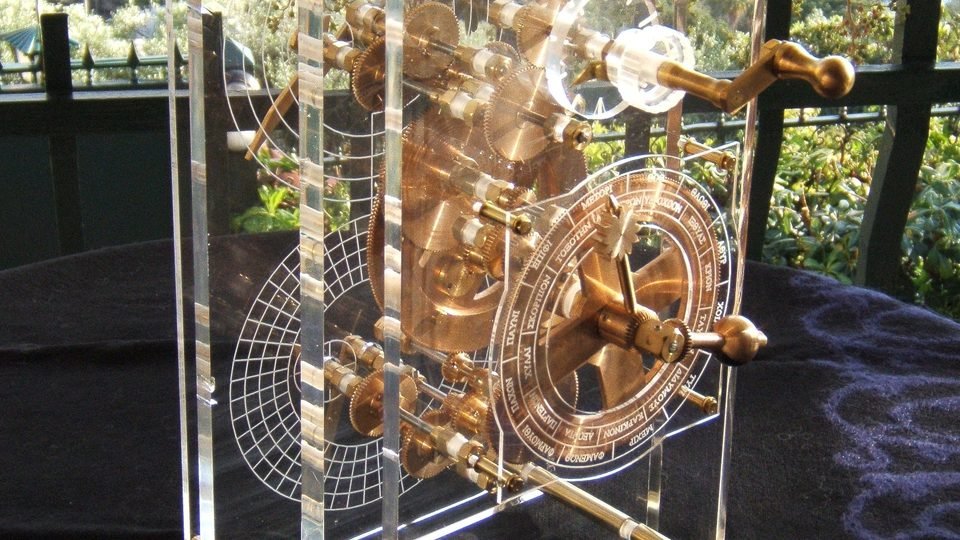 Modelo mecânico moderno do mecanismo de Anticítera, em caixa transparente: engrenagens de bronze à mostra e, na frente, o mostrador com os nomes dos meses em grego