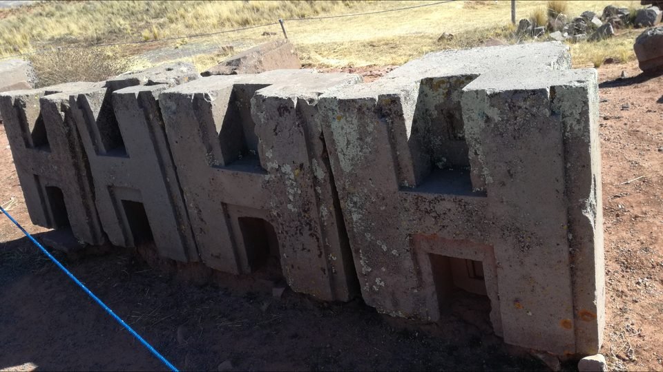 Fileira de blocos de andesito cinzento com perfil em forma de H, alinhados lado a lado sobre terra avermelhada em Puma Punku