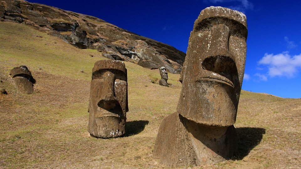 Moais na encosta de Rano Raraku, na Ilha de Páscoa, enterrados até o peito na grama, com o paredão da pedreira ao fundo sob céu azul