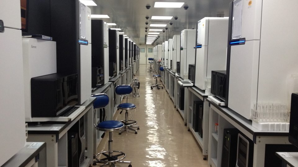 Corredor longo de um laboratório de sequenciamento de DNA, com fileiras de sequenciadores brancos dos dois lados e bancos azuis vazios