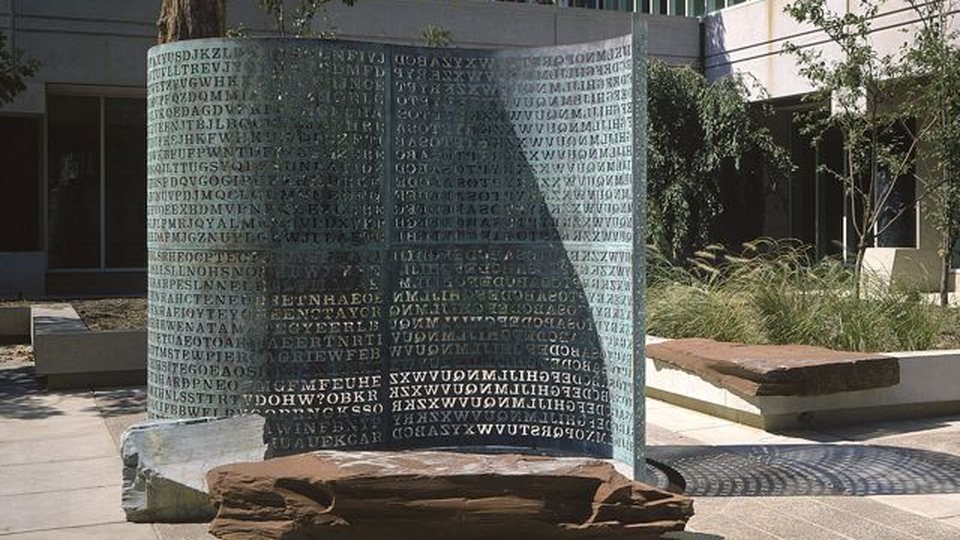 A escultura Kryptos no pátio da sede da CIA: uma lâmina curva de cobre esverdeado toda recortada em letras, sobre uma laje de pedra avermelhada