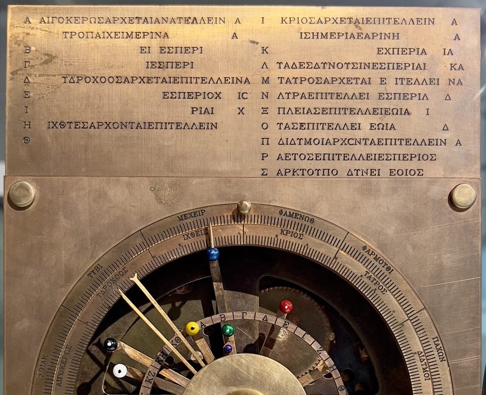 Réplica moderna do mecanismo de Anticítera: placa de latão com duas colunas de letras gregas gravadas no alto e, embaixo, a parte de cima do mostrador da frente, com anéis graduados e ponteiros