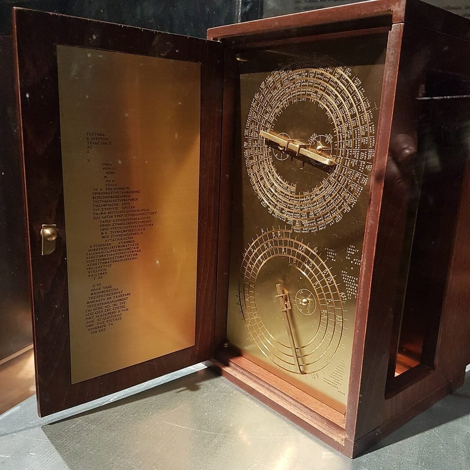Réplica moderna do mecanismo em caixa de madeira com placa de latão dourado: à direita, os dois grandes mostradores em espiral; na porta aberta, texto em grego