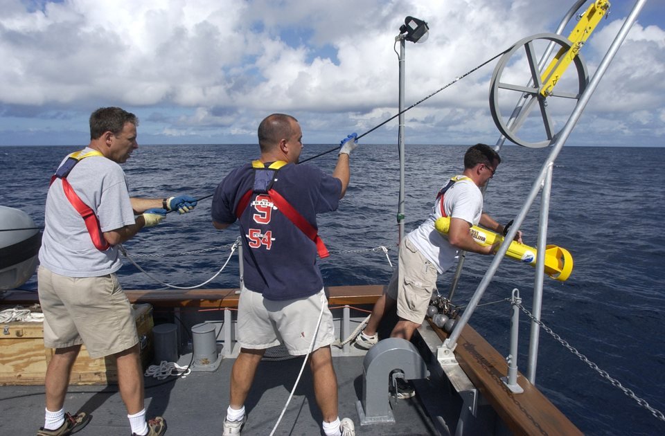 Três homens no convés de popa de um navio puxam o cabo de um sonar de varredura lateral, um cilindro amarelo com aletas, recolhido da água em mar aberto
