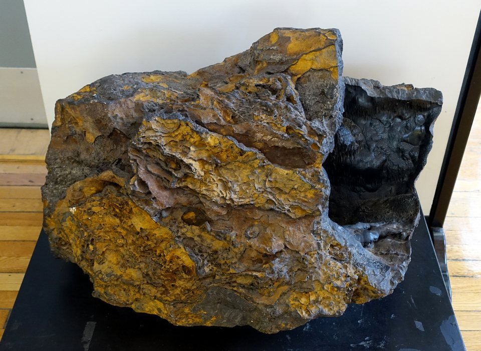Grande pedaço de limonita, minério de ferro de pântano, em exposição de museu: rocha escura e porosa com manchas e crostas amarelo-ocre