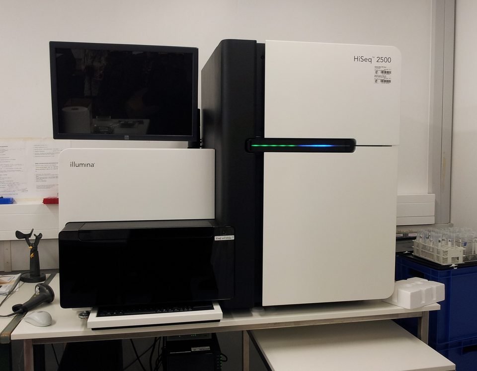 Sequenciador de DNA de bancada, um aparelho branco e preto do tamanho de um armário pequeno, com monitor ao lado, num laboratório