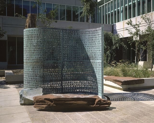 A escultura Kryptos no pátio da sede da CIA: uma lâmina vertical de cobre esverdeado, curva em S, toda recortada em letras, apoiada sobre uma laje de pedra avermelhada, com um tronco de madeira petrificada atrás e prédios de vidro ao fundo