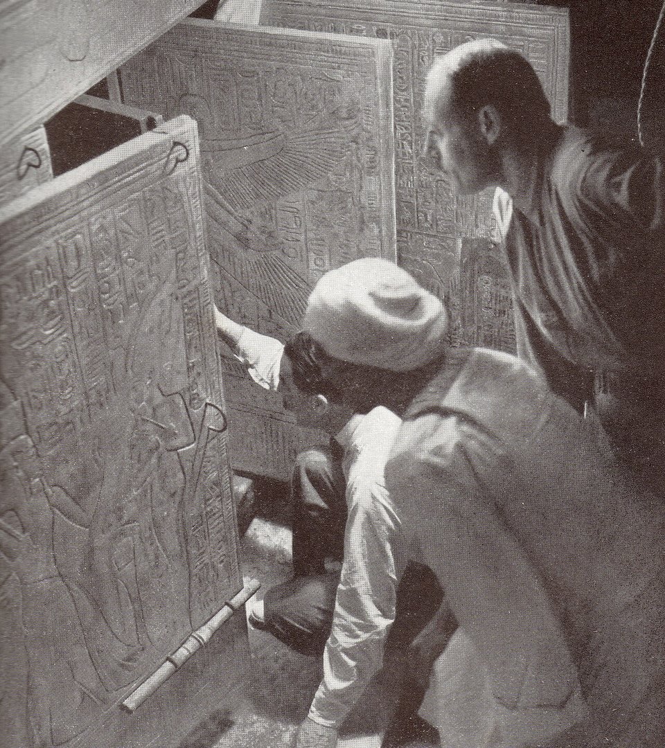 Fotografia em preto e branco: Howard Carter, ajoelhado, abre as portas de um santuário dourado coberto de hieróglifos dentro do túmulo de Tutancâmon, observado por dois homens