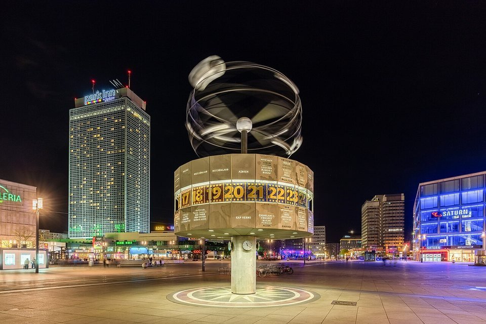 O relógio mundial da Alexanderplatz, em Berlim, à noite: uma coluna com um cilindro de painéis com nomes de cidades e as horas, encimado por uma estrutura metálica de órbitas em movimento