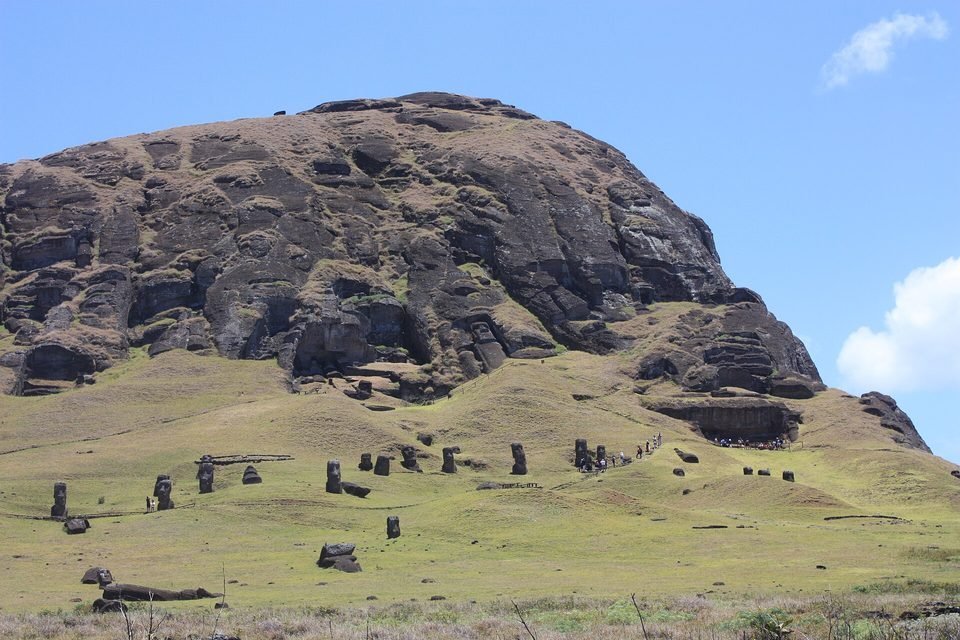 The slope of the Rano Raraku volcano under a blue sky: the dark rock face at the top, cut with quarrying niches, and on the grassy slope dozens of standing moai, with only head and shoulders above the ground