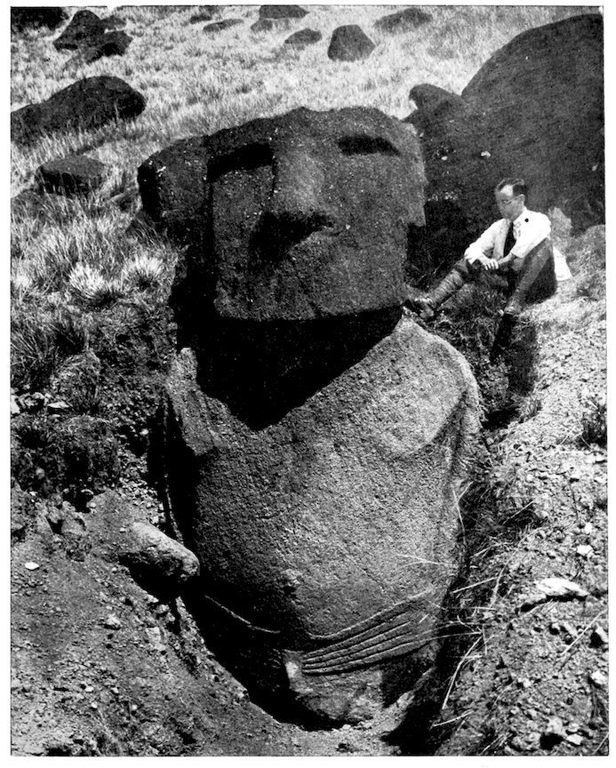 Black-and-white photograph: a Rano Raraku moai excavated in a deep trench, with its body and hands exposed below ground level; a man in a suit sits on the edge of the trench, beside the head