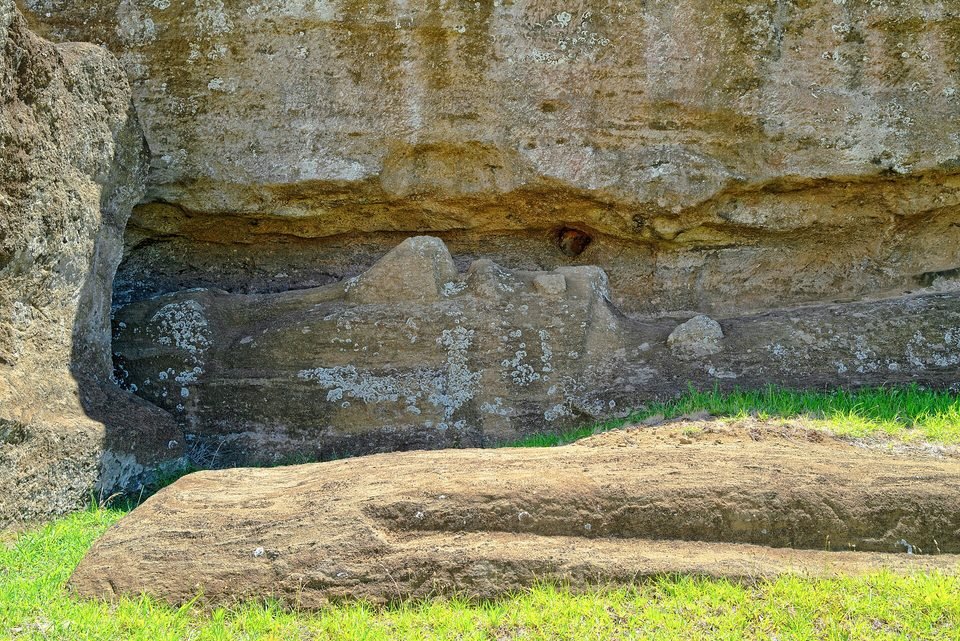 Unfinished moai lying in the rock of the Rano Raraku quarry: the profile of the face, with nose and chin, carved sideways into a ledge of the rock face, still attached to the stone, with another worked block in the foreground on the grass