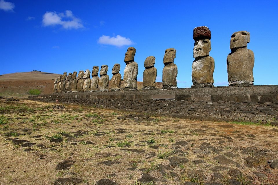 Ahu Tongariki: a row of fifteen standing moai on a long stone platform, backs to the sea, one of them with the red stone cylinder on its head, under a blue sky