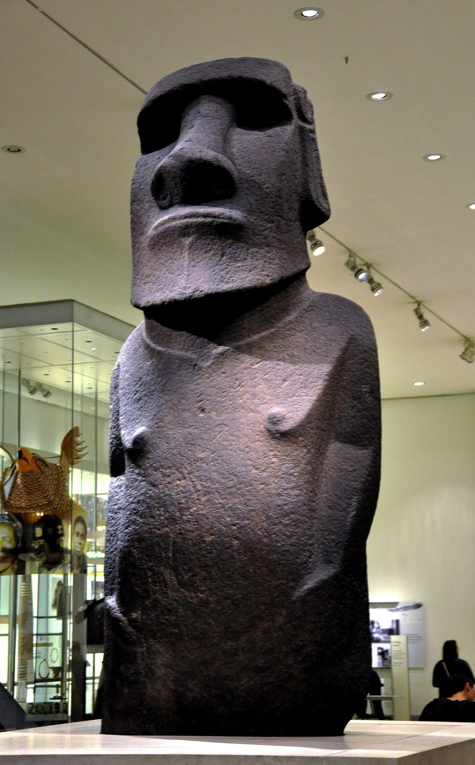 Hoa Hakananai'a, a dark basalt moai, displayed on a pedestal in a gallery of the British Museum, seen from below, with its elongated face and hands carved on the belly