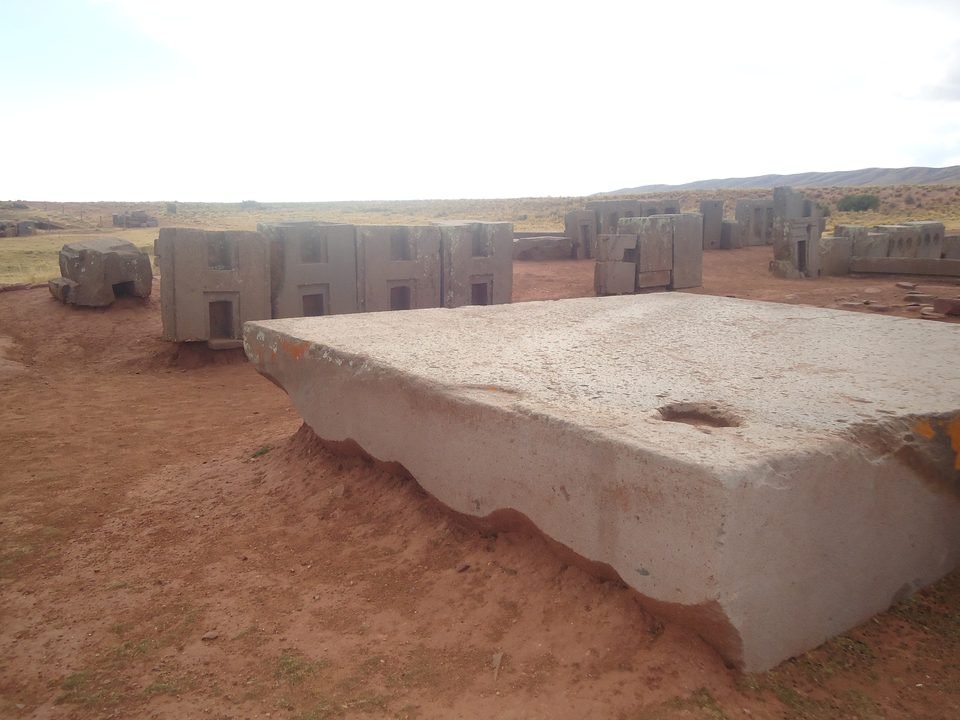 Large reddish sandstone slab, flat on top with an irregular edge, lying on red earth; in the background, rows of H-shaped andesite blocks