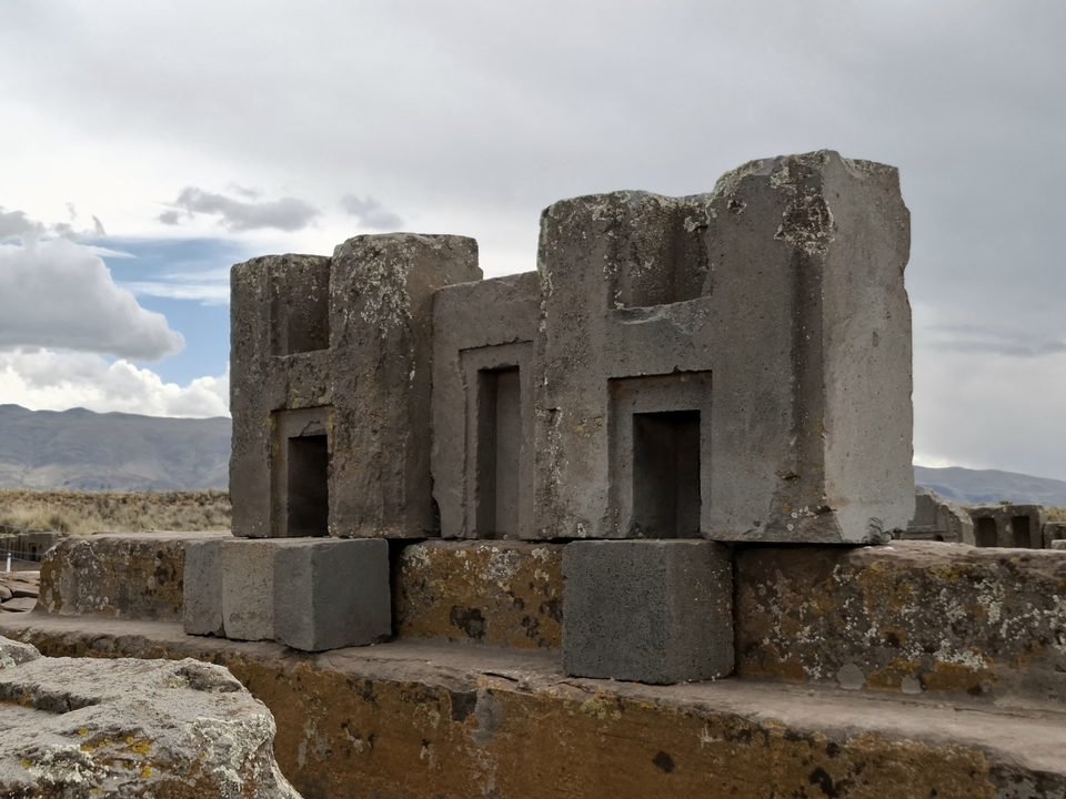 Two grey andesite blocks with an H-shaped profile, with niches and right-angled inner corners, set on smaller blocks and a sandstone base, under a cloudy sky