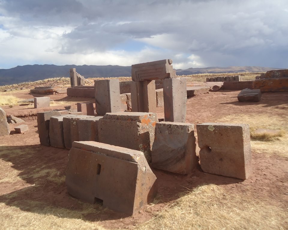 Grey andesite and reddish sandstone blocks scattered over red earth at Puma Punku, some stacked, with a fragmented doorway in the background and mountains on the horizon