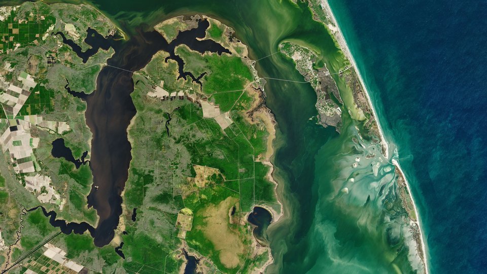 Imagem de satélite em cores naturais: no alto, à direita, a ilha de Roanoke com pontes ligando-a ao continente e ao cordão de areia; à direita, o oceano Atlântico; embaixo, a barra de Oregon Inlet com bancos de areia claros