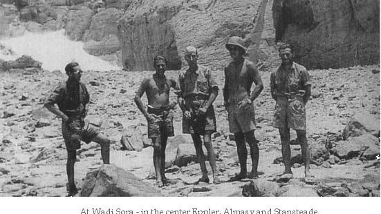 Fotografia em preto e branco de cinco homens de pé sobre pedregulhos junto a um paredão rochoso no Wadi Sura